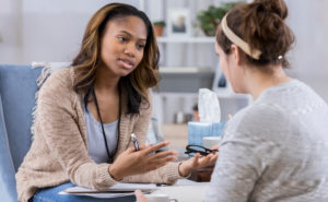 Serious female counselor gestures while talking with Caucasian female client. The counselor is holding eyeglasses and a pen. They are discussing serious issues.
