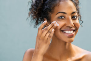 Beautiful african american woman with smooth skin applying moisturizer face cream to her cheek. Beauty young woman taking care of skin. Happy girl applying cosmetic moisturiser treatment isolated on background and looking at camera with copy space.